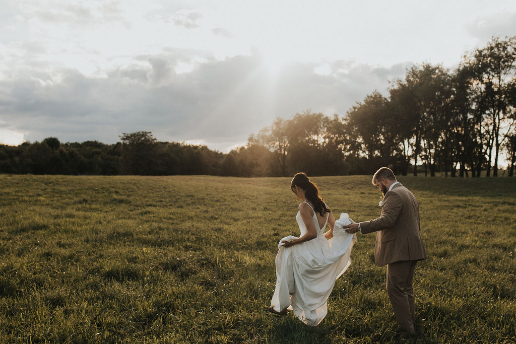 groom carries bride's wedding dress train in field