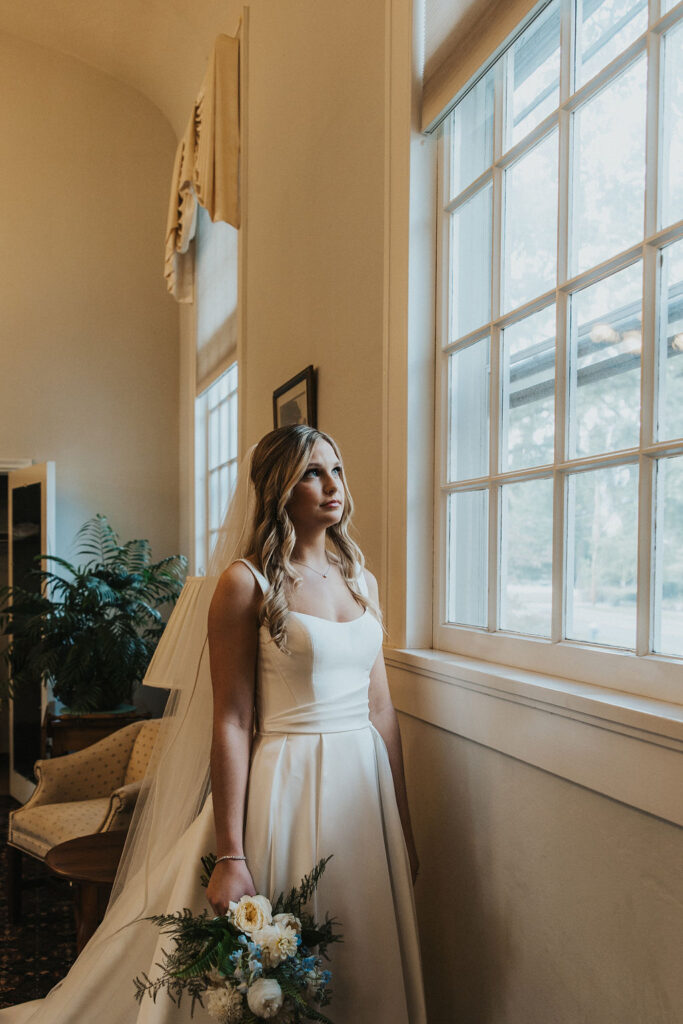 bride stands beside window with wedding bouquet 