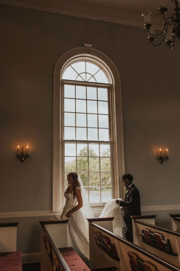 groom carries bride's veil beside church window